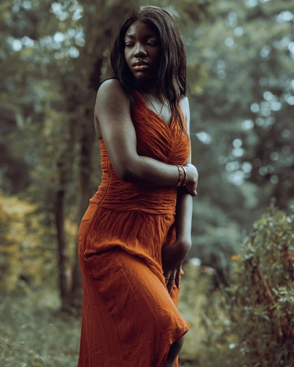 a woman in an orange dress standing in the woods, inspired by Elsa Bleda, pexels contest winner, renaissance, dark brown skin, muted brown, doing a sassy pose, low quality photo