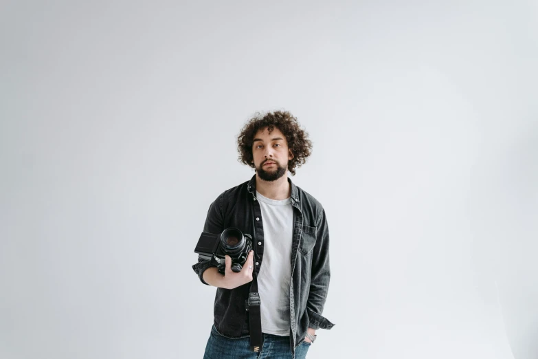 a man standing in front of a white backdrop holding a camera, scruffy man, daniel mirante, ash thorp, plain background