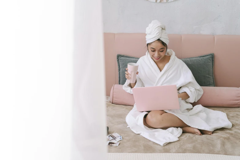 a woman sitting on a bed using a laptop, trending on pexels, happening, pastel pink robes, skincare, milk and mocha style, white sarong