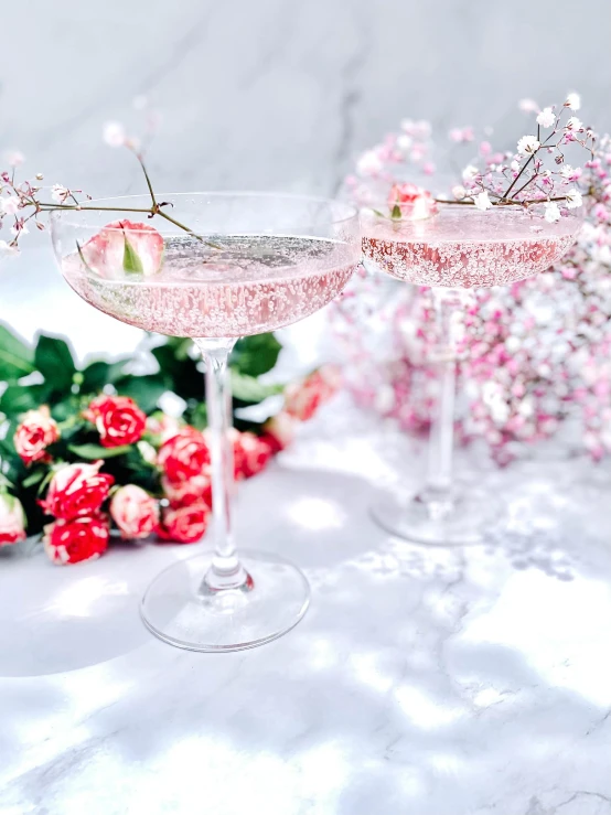 a couple of wine glasses sitting on top of a table, pink flowers, with a drink, sparkels, promo image