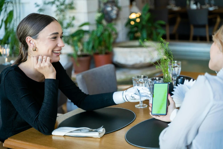 two women sitting at a table talking to each other, pexels contest winner, happening, holding a very advance phone, avatar image, close - up photo, green