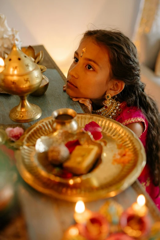 a little girl sitting at a table with a plate of food, hindu aesthetic, emitting light ornaments, ( ( theatrical ) ), 24k