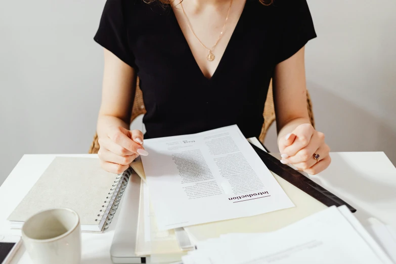 a woman sitting at a table with papers and a pen, an album cover, by Julia Pishtar, pexels contest winner, wearing business casual dress, she is wearing a black tank top, wearing elegant jewellery, holding notebook