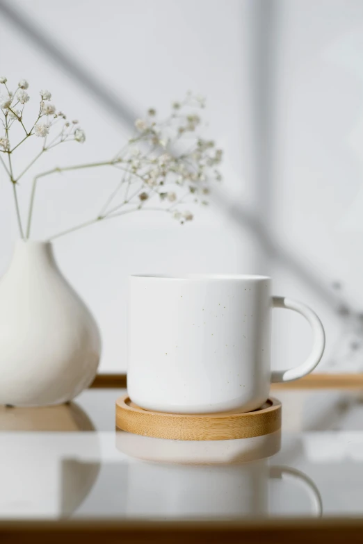 a white cup sitting on top of a table next to a vase, a picture, textured base ; product photos, made of bamboo, with a white mug, detail shot