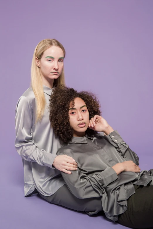 two women sitting next to each other on a purple background, trending on pexels, antipodeans, attractive androgynous humanoid, grey clothes, arm around her neck, mixed race