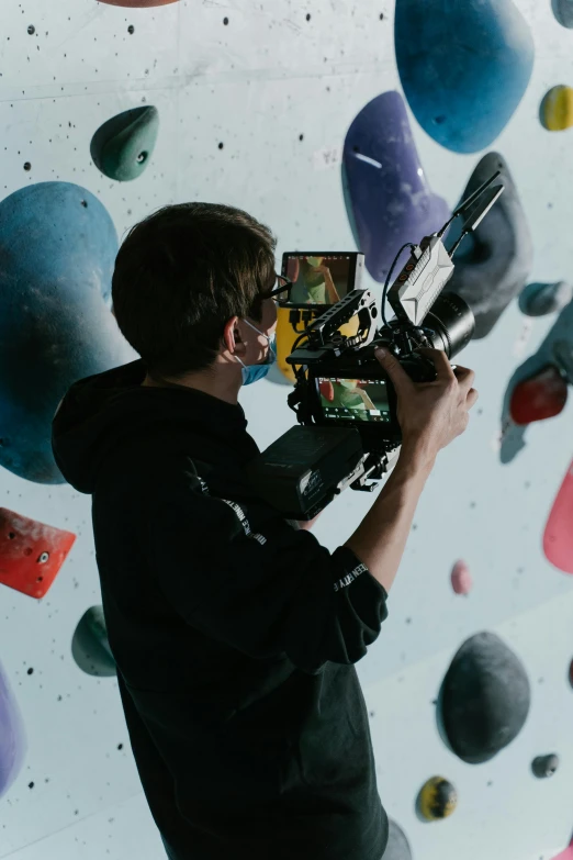 a man holding a video camera in front of a climbing wall, unsplash, video art, film studio, adam ondra, vhs recording, looking across the shoulder