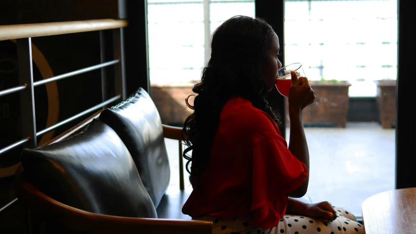 a woman sitting at a table drinking from a cup, pexels contest winner, dark skinned, lounge, from the back, crimson