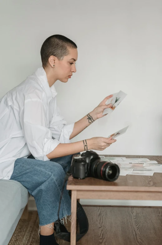 a man sitting on a couch holding papers and a camera, a picture, by Pablo Rey, on a white table, an epic non - binary model, inspect in inventory image, seated at a table