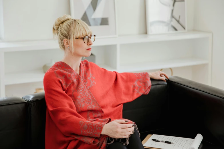 a woman sitting on a couch in a living room, by Lisa Milroy, pexels contest winner, red cape, office clothes, with glasses on, blonde swedish woman