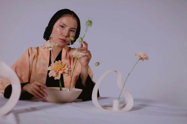 a woman sitting at a table with a bowl of flowers, an album cover, inspired by Kim Tschang Yeul, slow motion fashion, video still, on a pale background, still frame