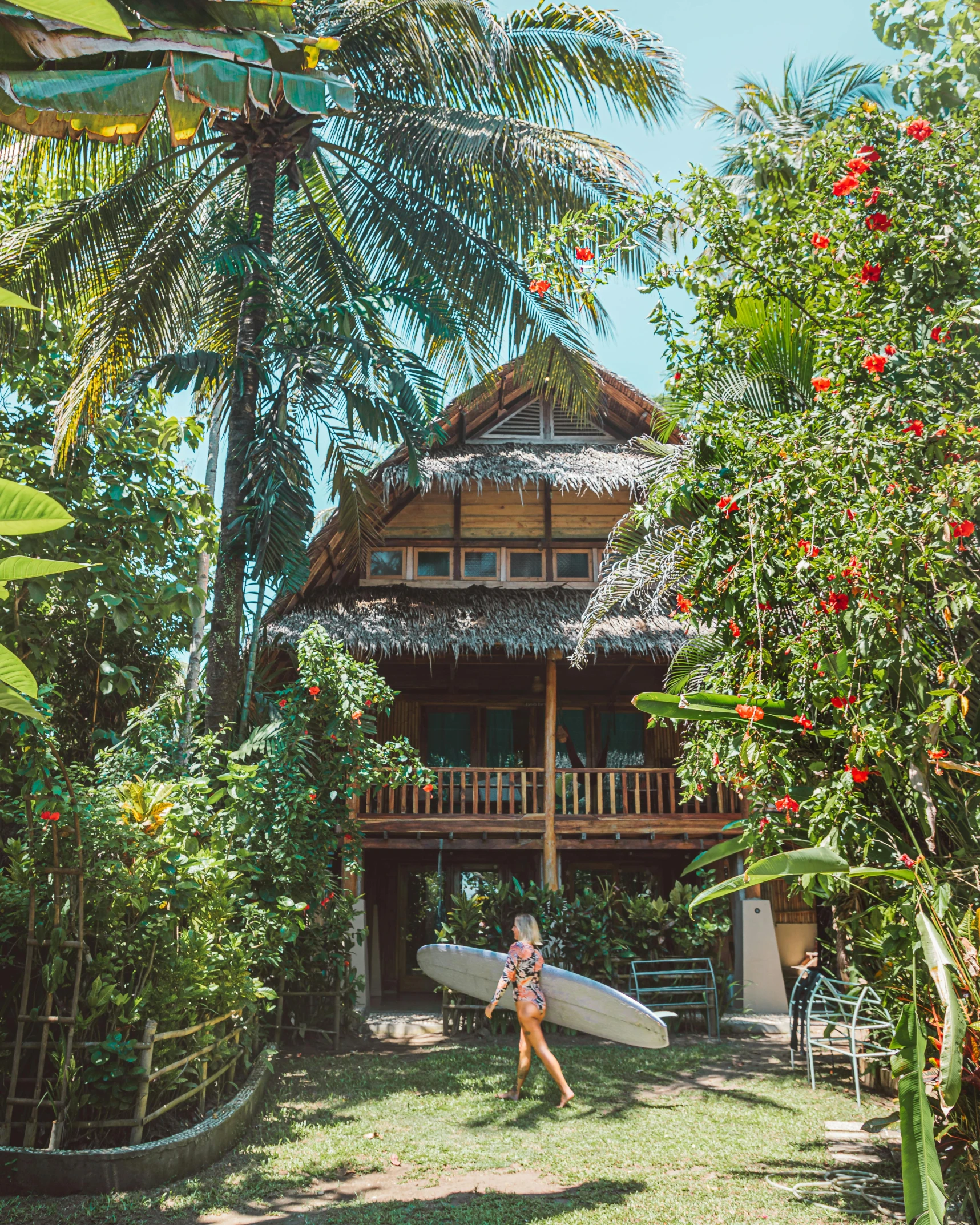 a woman carrying a surfboard on top of a lush green field, by Sam Dillemans, sumatraism, wooden cottage, with fruit trees, full building, flatlay