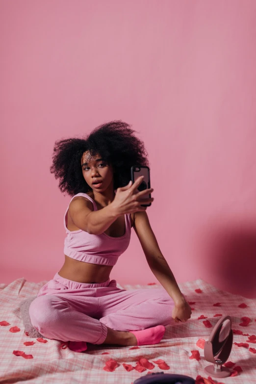 a woman sitting on top of a bed holding a cell phone, by Lily Delissa Joseph, trending on pexels, happening, pink background, wearing a tracksuit, natural hair, taking selfies