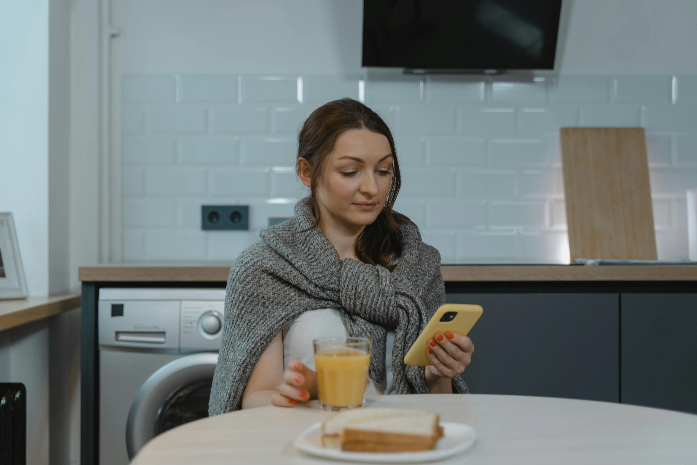 a woman sitting at a kitchen table using a cell phone, by Adam Marczyński, trending on pexels, realism, grey, wearing a sweater, pregnancy, breakfast