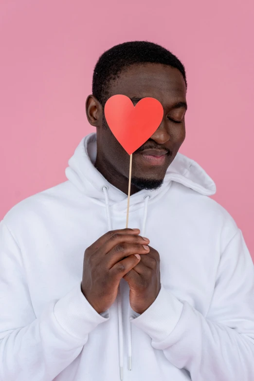 a man in a white hoodie holding a red heart on a stick, trending on pexels, with brown skin, wearing a pastel pink hoodie, promo image, essence