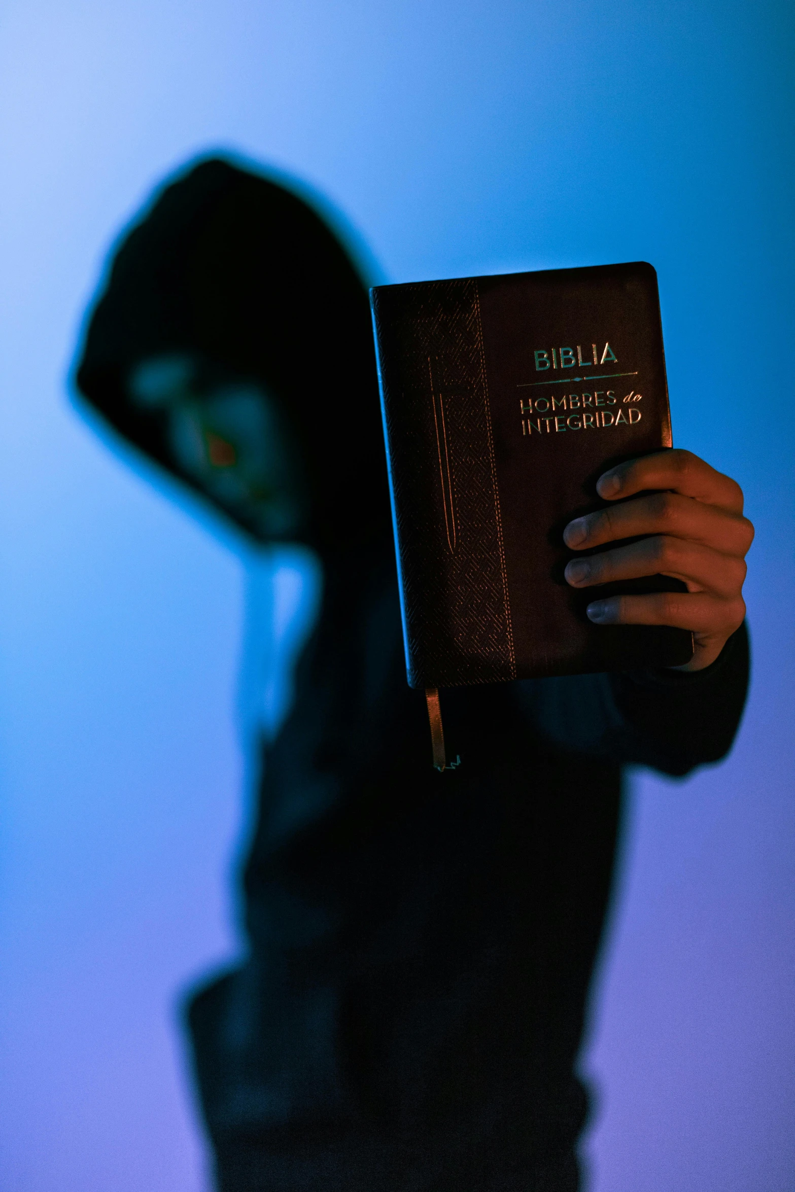 a person holding a book in front of a blue background, unilalianism, wearing a dark hood, bible, evil glow, masked person in corner