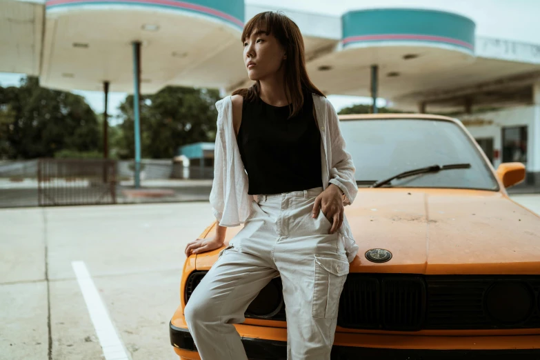 a woman sitting on the hood of a car, inspired by Gang Hui-an, pexels contest winner, white pants, set on singaporean aesthetic, in front of a garage, with two front pockets