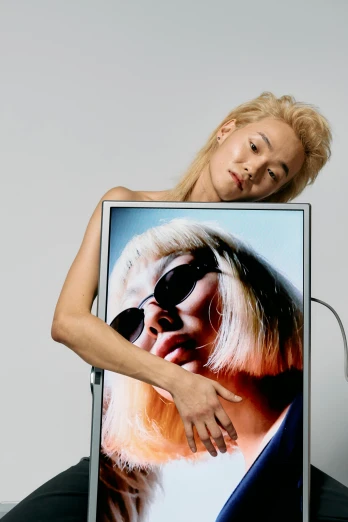 a woman sitting on a chair holding a picture of herself, an album cover, inspired by Nan Goldin, photorealism, platinum blonde, sun-hyuk kim, in front of a computer, a portrait of issey miyake
