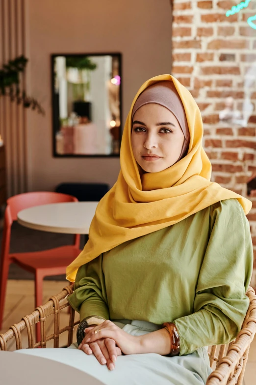 a woman sitting in a chair wearing a yellow scarf, inspired by Nazmi Ziya Güran, trending on pexels, hurufiyya, standing in a restaurant, square, proud serious expression, islamic