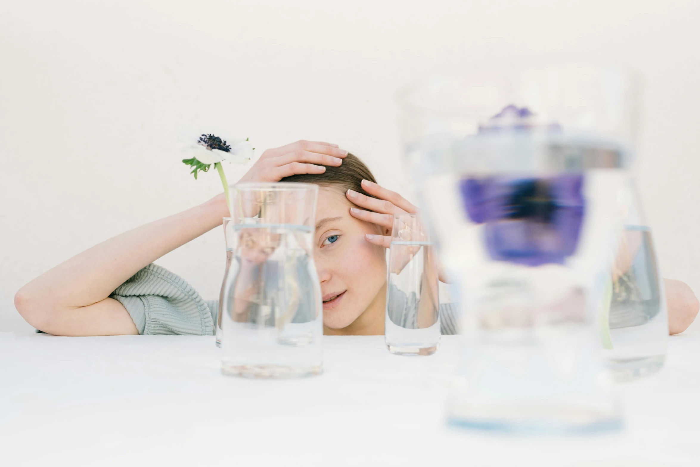 a woman sitting at a table with a flower in her hair, a still life, inspired by Méret Oppenheim, unsplash, lsd water, on a white table, person made out of glass, looking tired