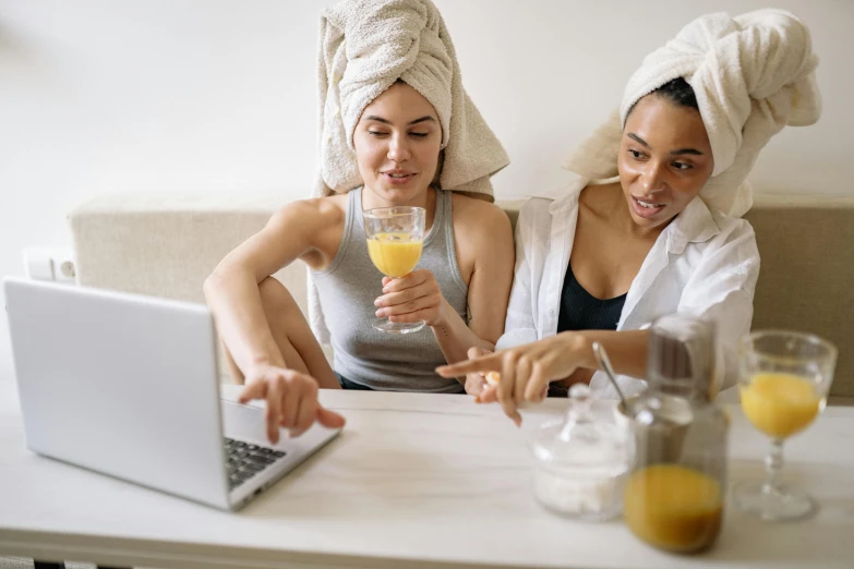 two women sitting at a table with a laptop and a glass of orange juice, trending on pexels, happening, wearing a towel, without makeup, dirty linen robes, people watching around