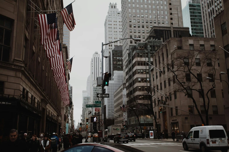 a car driving down a city street next to tall buildings, a photo, pexels contest winner, american flags, background image, people walking around, gray skies