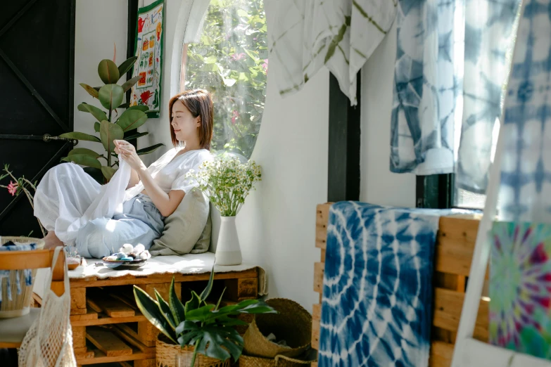 a woman sitting on top of a wooden bench next to a window, trending on pexels, arts and crafts movement, fabrics and textiles, cats and plants, relaxing on a couch, wearing a tie-dye t-shirt