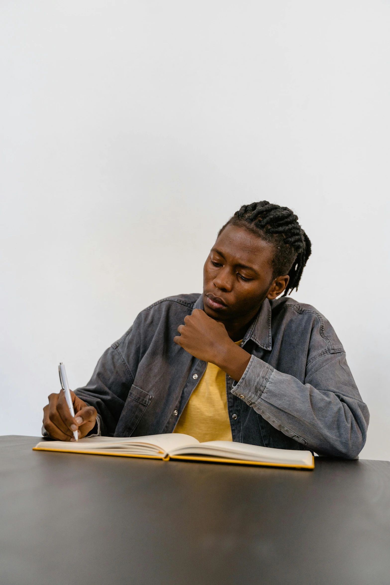 a man sitting at a table writing in a book, by Stokely Webster, black teenage boy, empty background, telling stories, ignant