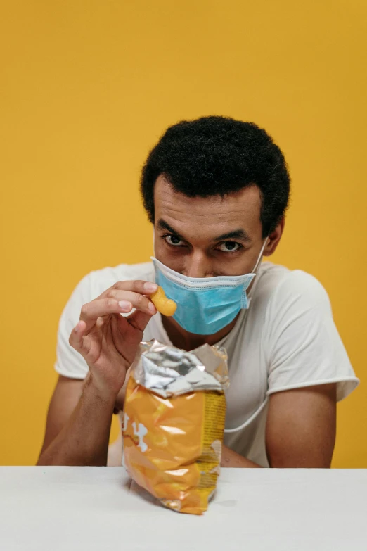 a man sitting at a table with a bag of food in front of him, a picture, yellow backdrop, surgical mask covering mouth, quack medicine, about to consume you