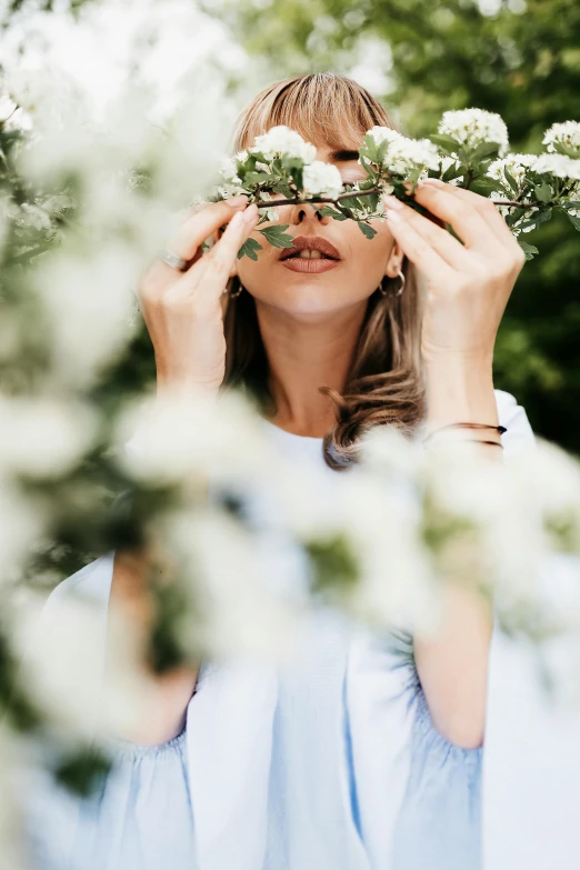 a woman holding a flower in front of her face, by Zofia Stryjenska, trending on unsplash, white blossoms, with soft bushes, hiding behind obstacles, woman made of plants