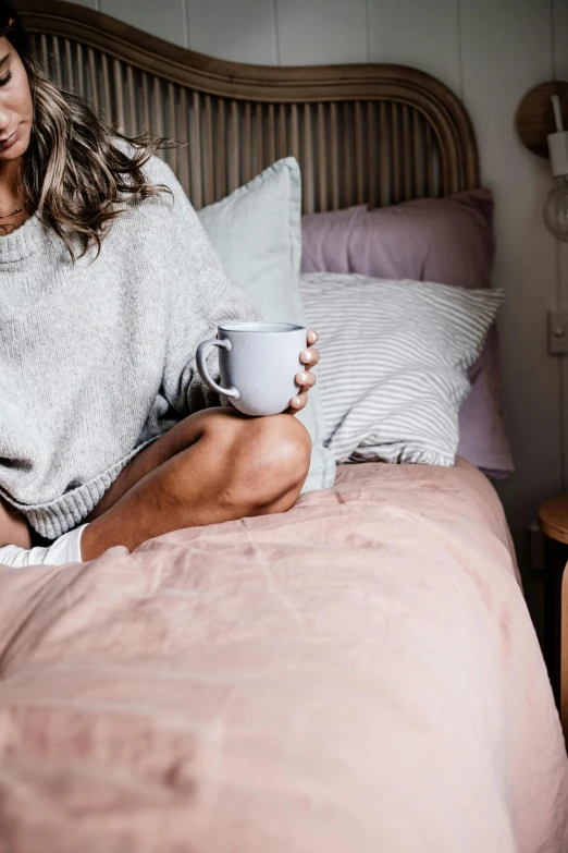 a woman sitting on a bed with a cup of coffee, happening, manuka, light grey, pink, chilly