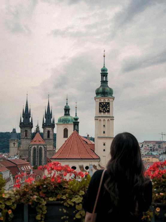 a woman standing in front of a clock tower, by Julia Pishtar, pexels contest winner, art nouveau, prague in the background, panorama view, black domes and spires, flowers in foreground