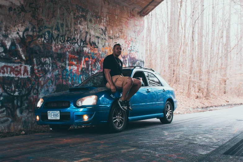 a man sitting on the hood of a blue car, a portrait, inspired by Michael Gustavius Payne, pexels contest winner, graffiti, subaru, an olive skinned, teddy fresh, sitting under bridge