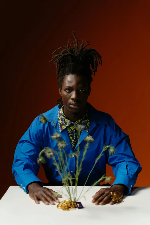 a man with dreadlocks sitting at a table, an album cover, inspired by Charles Martin, wearing blue jacket, adut akech, flowers, portrait n - 9