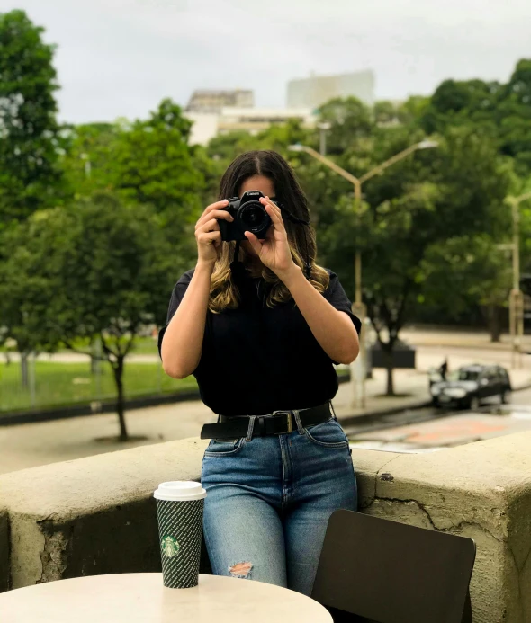 a woman sitting at a table taking a picture, wearing a dark shirt and jeans, são paulo, profile image, trees in the background