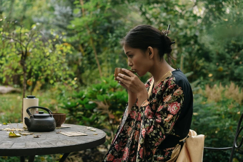 a woman sitting at a table with a cup of coffee, inspired by Itō Shinsui, unsplash, shin hanga, background: assam tea garden, the movie, wearing floral chiton, tessa thompson