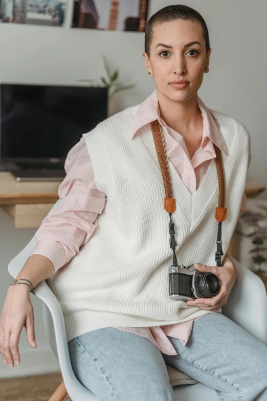 a woman sitting in a chair holding a camera, wearing a white sweater, wearing a vest, wearing a white button up shirt, leather jewelry