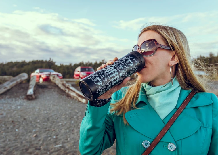 a woman in a blue coat drinking from a bottle, by Nina Hamnett, pexels contest winner, solar eclipse in iceland, monster energy drink, drink more coffee, near the beach