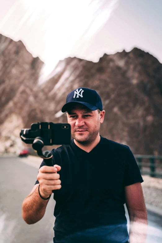 a man standing on the side of a road holding a camera, standing in front of a mountain, tiktok video, jony ive, avatar image