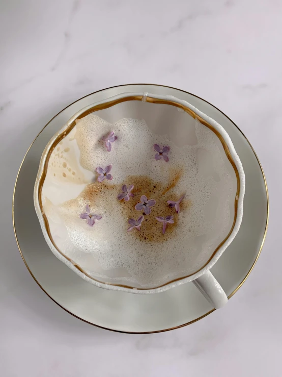 a close up of a cup of coffee on a table, inspired by Méret Oppenheim, lavender flowers, disintegrating, white with gold accents, brooke ashling