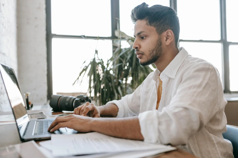 a man sitting at a desk using a laptop computer, a screenshot, pexels contest winner, aboriginal australian hipster, wearing a white button up shirt, concentration, thumbnail