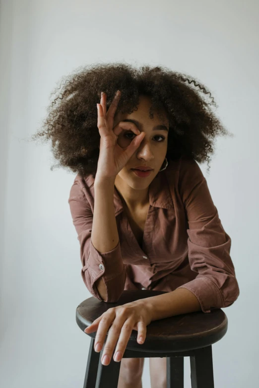 a woman sitting on top of a wooden stool, by Lily Delissa Joseph, pexels contest winner, (dark shorter curly hair), greeting hand on head, ( brown skin ), portrait androgynous girl