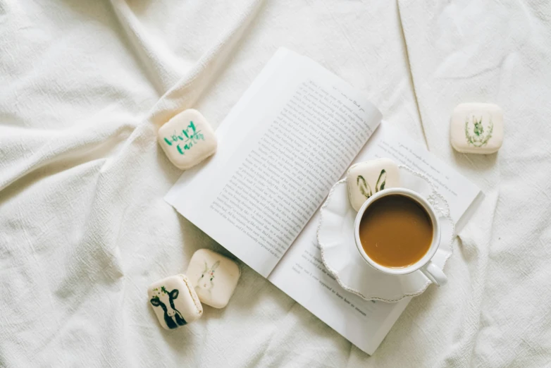 an open book sitting on top of a bed next to a cup of coffee, by Emma Andijewska, trending on unsplash, romanticism, marshmallows, on a white table, laying back on a pillow, thumbnail