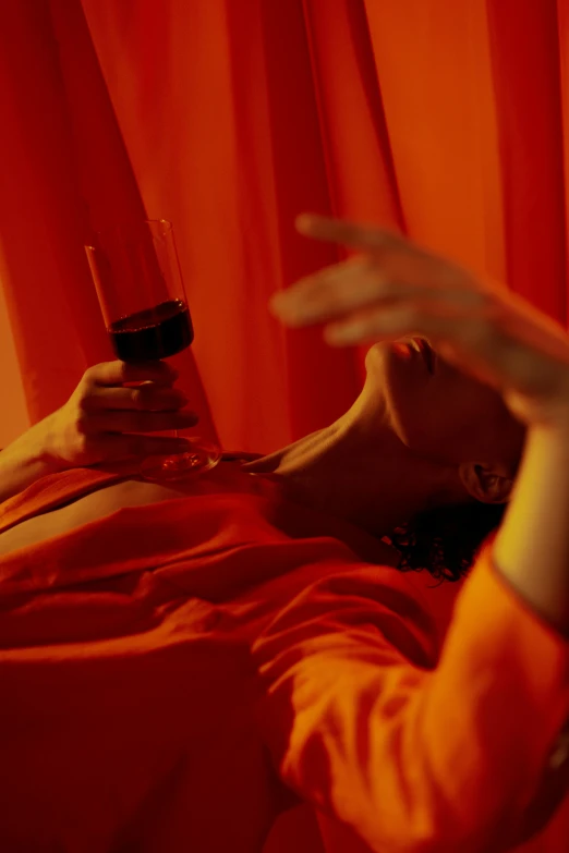 a woman laying on a bed holding a cell phone, inspired by Nan Goldin, renaissance, enjoying a glass of wine, vibrant orange, showstudio, red curtain