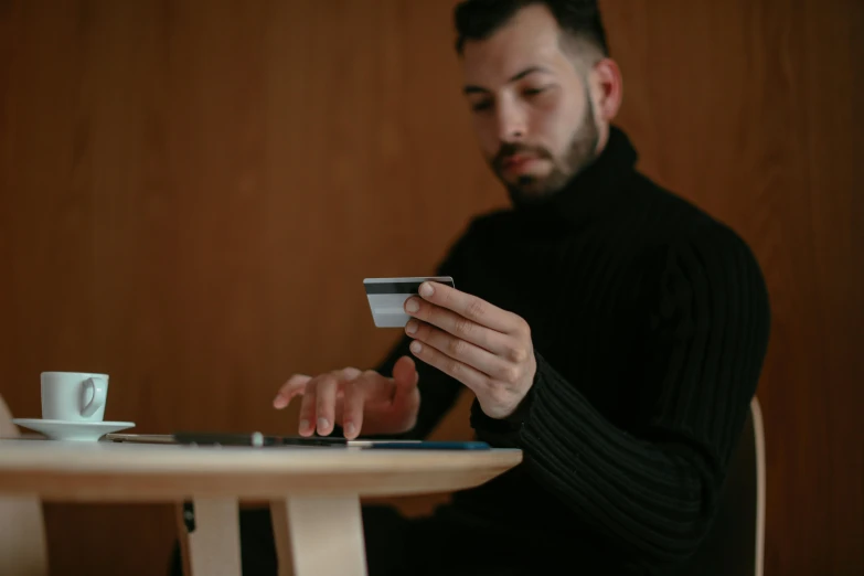 a man sitting at a table using a cell phone, ecommerce photograph, wearing a black sweater, focus on card, avatar image