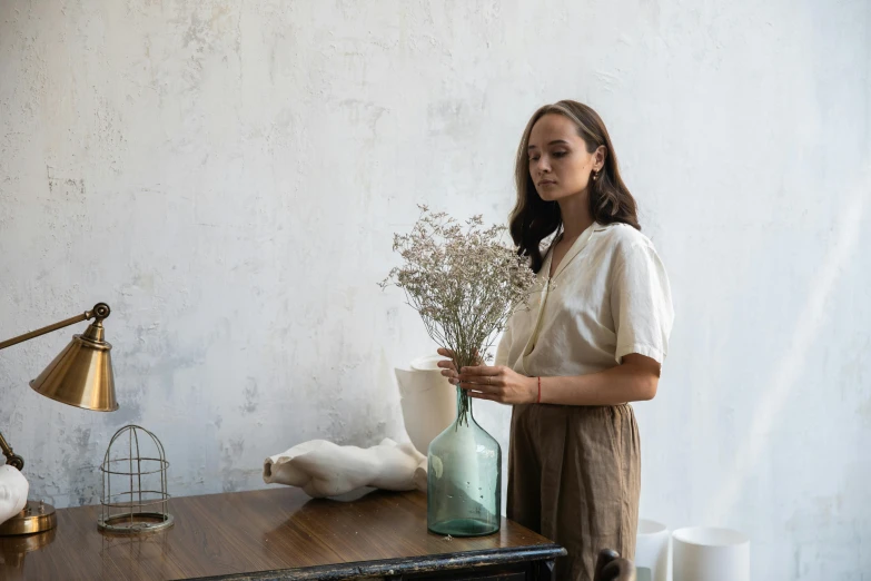 a woman standing next to a table with a vase of flowers, inspired by Camille Corot, featured on unsplash, wearing a linen shirt, facing left, sustainable materials, holding a bottle of arak