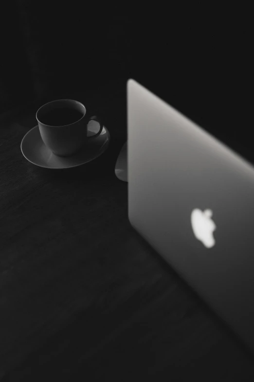 a laptop computer sitting on top of a table next to a cup of coffee, a black and white photo, by Byron Galvez, pexels, postminimalism, apple logo, dark dance photography aesthetic, black silhouette, tea