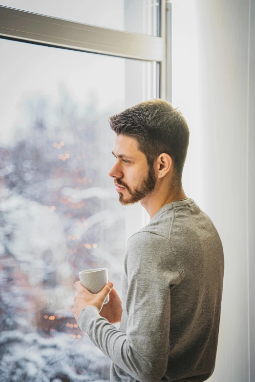 a man standing in front of a window holding a cup of coffee, trending on pexels, romanticism, looking off to the side, gray men, young spanish man, : :