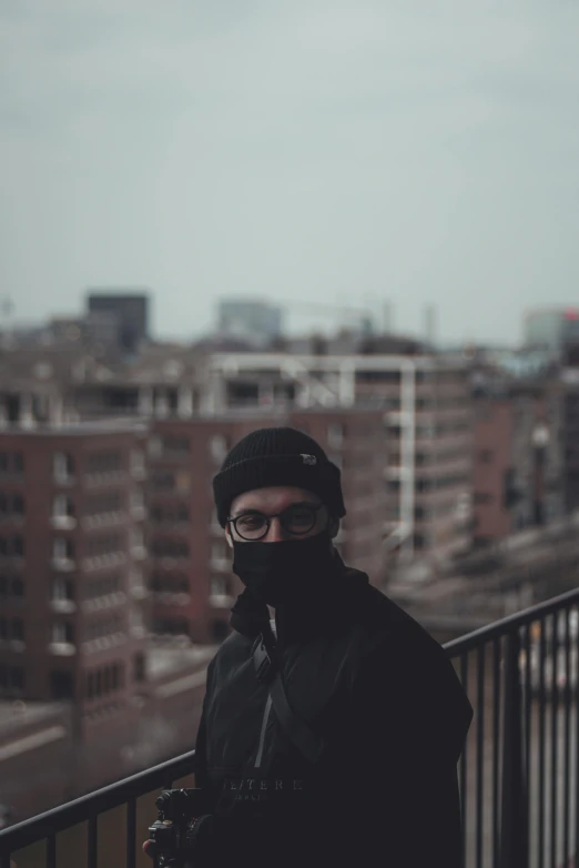 a man standing on a balcony with a camera, by Thomas Wijck, unsplash, wearing all black mempo mask, staring into the camera, bleak color, city in the distance