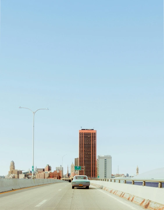 a car driving down a highway with tall buildings in the background, a photo, inspired by Robert Bechtle, photorealism, rhode island, clemens ascher, tall bridge with city on top, shoreline