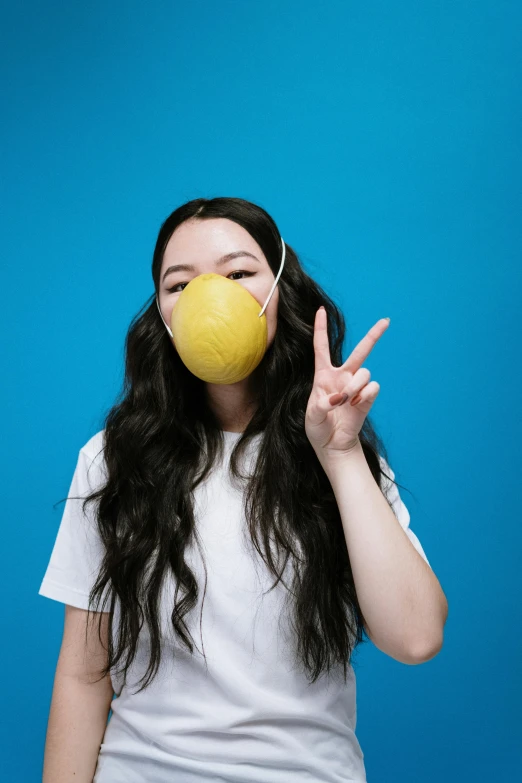 a woman holding an apple in front of her face, an album cover, trending on pexels, respirator, blue and yellow color theme, chinese girl, peace sign